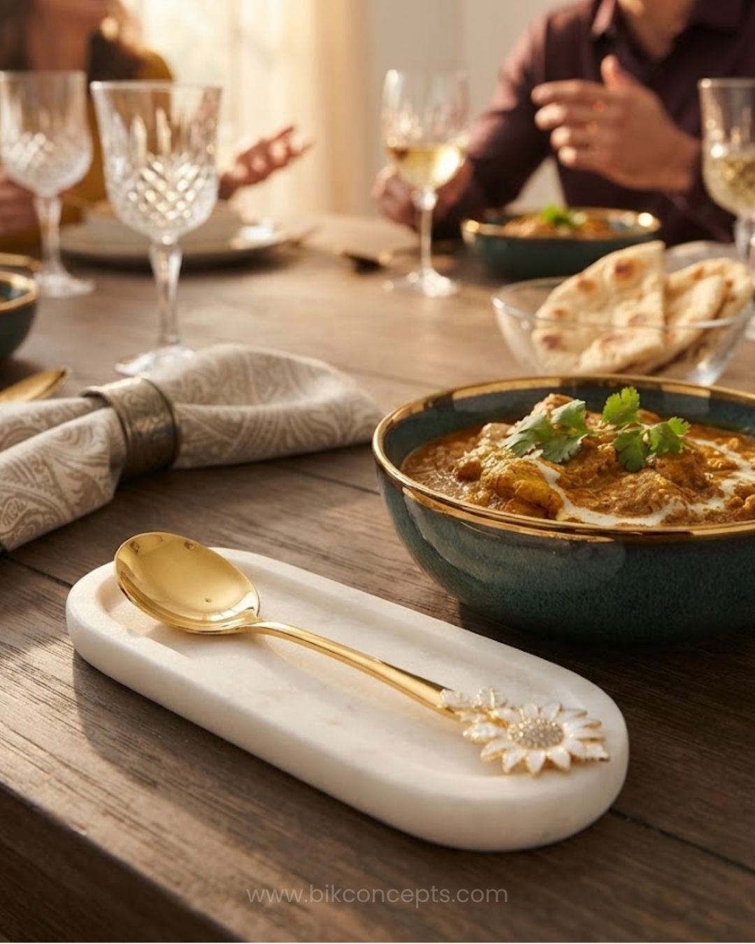 A close-up dining scene showing a white marble spoon rest with a gold spoon and a white flower detail, set on a wooden table beside a bowl of curry. A person is seated in the softly blurred background at the head of the table, with wine glasses and flatbread visible, Gold Stainless Steel Serving Spoon, Sunflower Decorative Serving Set, 11 Inch Round Gold Serving Spoons, Set of 3 Gold Buffet Spoons, Set of 5 Tableware Serving Spoons, Floral Enamel Handle Flatware, Luxury Gold Serving Utensils, Decorative Kit