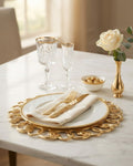 A styled table setting with a gold-plated charger plate, white dinner plate, cream napkin, gold cutlery, two crystal wine glasses with gold rims, a small bowl of chocolates, and a gold vase holding a white rose on a white marble table, Gold Charger Plates Set of 6, Interlocking Loop Plastic Charger Plates, 13 Inch Round Service Plates, Luxury Wedding Table Decor US, Catering Charger Plates Bulk, Openwork Chain Link Underplates, Gold Metallic Place Setting Base Plates, Modern Event Decor Charger Plates, Whol