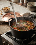 A black cast iron Dutch oven on a gas stove burner, filled with a hearty stew and a metal ladle resting inside. A copper lid sits on a cloth in the background, with bowls and spices Heavy Duty Stainless Steel 5 oz Ladle, Commercial Grade Soup Ladle 14.5 Inch, Thick Metal Portion Control Ladle, One Piece Stainless Steel Serving Ladle, Hotel Buffet Soup Ladle with Hook, Professional Catering Gravy Ladle 5 oz, Large Stainless Steel Ladle for Stockpot, Restaurant Quality Metal Sauce Dipper, Premium Silver Servi