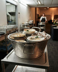 A large stainless steel buffet tub filled with dirty plates, bowls, napkins, and cutlery, placed on a metal cart in a restaurant or catering kitchen. Commercial Stainless Steel Utility Tub, Large Stainless Steel Beverage Tub, 23 Inch Stainless Steel Prep Basin, Hotel Catering Ice Bucket Tub, Heavy Duty Stainless Steel Wash Tub, Commercial Kitchen Food Prep Bowl, Large Party Beverage Chiller Tub, Stainless Steel Storage Tub with Handles, Polished Silver Catering Basin, Multipurpose Commercial Kitchen Tub