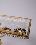 A close-up of a decorative clear glass tray with an ornate gold metal frame, featuring a gold leaf motif and a central gold flower embellishment with a clear rhinestone.