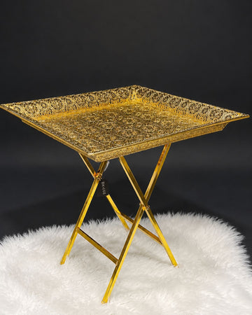 A square gold metal serving tray with an ornate embossed pattern, supported by a matching gold folding metal stand,Golden Square Serving Table, Embossed Metal Square Tray Table, Luxury Gold Accent Side Table, Moroccan Style Gold Tea Table, Designer Folding Drink Table USA, Filigree Metal End Table, Hollywood Regency Gold Furniture, Luxury Home Hosting Accessories, Decorative Square Metal Tray Table 21 Inch, High End Gold Bar Cart Alternative