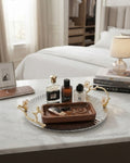 A styled bedroom scene with a round silver tray featuring gold-toned branch handles, holding a smaller brown wooden tray with jewelry and several perfume bottles. The setting includes a bed, lamp, books, and a marble tabletop, 12 Inch Stainless Steel Serving Tray, Luxury Jasmine Tray with Handles, High End Botanical Platter, Metal Serving Platter for Catering, Fluted Silver Cocktail Tray, Premium Catering Food Presentation USA, Designer Vanity Display Dish, Gold and Silver White Flower Tray
