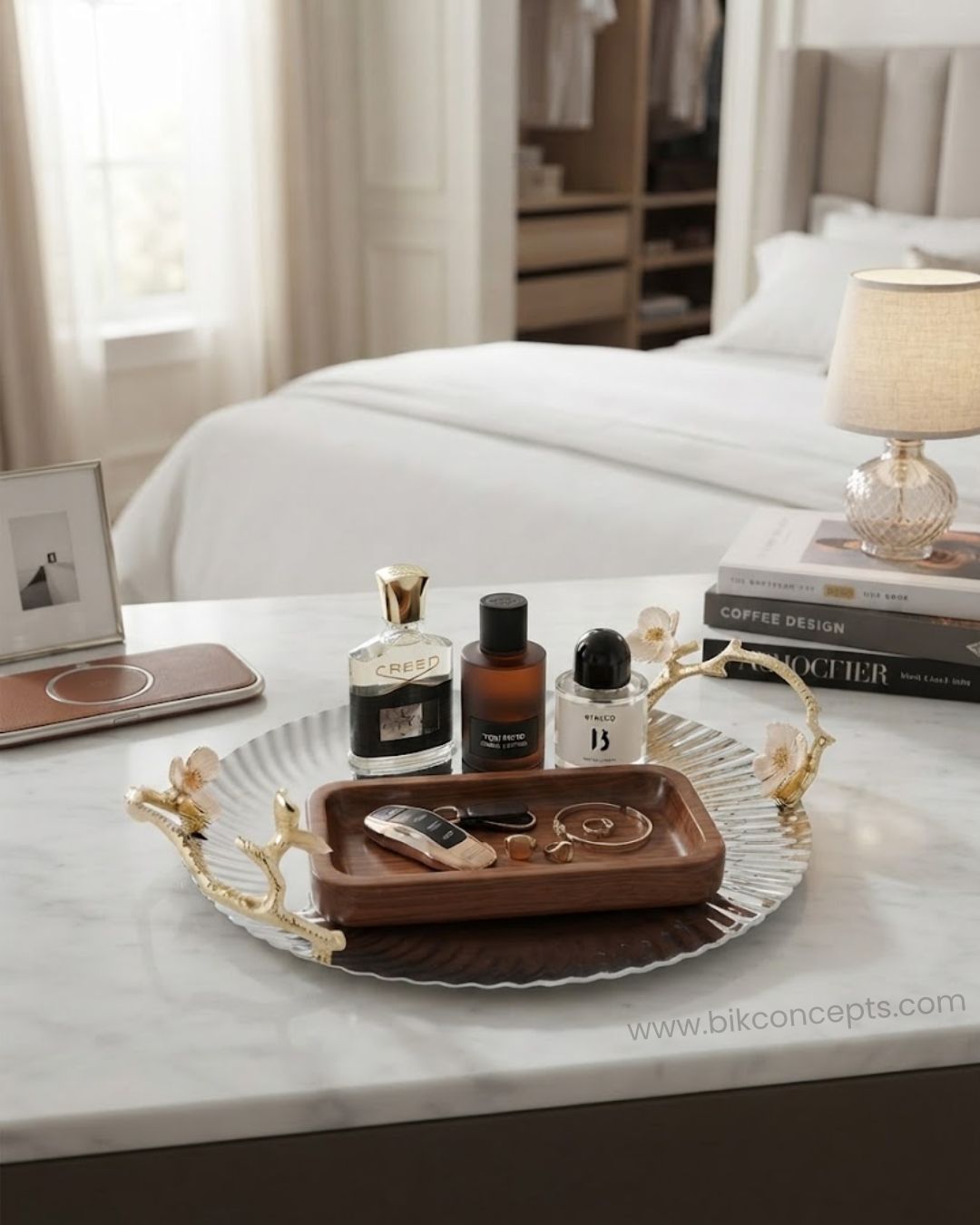 A styled bedroom scene with a round silver tray featuring gold-toned branch handles, holding a smaller brown wooden tray with jewelry and several perfume bottles. The setting includes a bed, lamp, books, and a marble tabletop, 12 Inch Stainless Steel Serving Tray, Luxury Jasmine Tray with Handles, High End Botanical Platter, Metal Serving Platter for Catering, Fluted Silver Cocktail Tray, Premium Catering Food Presentation USA, Designer Vanity Display Dish, Gold and Silver White Flower Tray