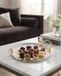 A round silver serving platter with gold-toned handles and floral accents, holding assorted chocolate-covered treats, placed on a white marble coffee table in a softly lit living room, 12 Inch Stainless Steel Serving Tray, Luxury Jasmine Tray with Handles, High End Botanical Platter, Metal Serving Platter for Catering, Fluted Silver Cocktail Tray, Premium Catering Food Presentation USA, Designer Vanity Display Dish, Gold and Silver White Flower Tray, Luxury Wedding Cateringware