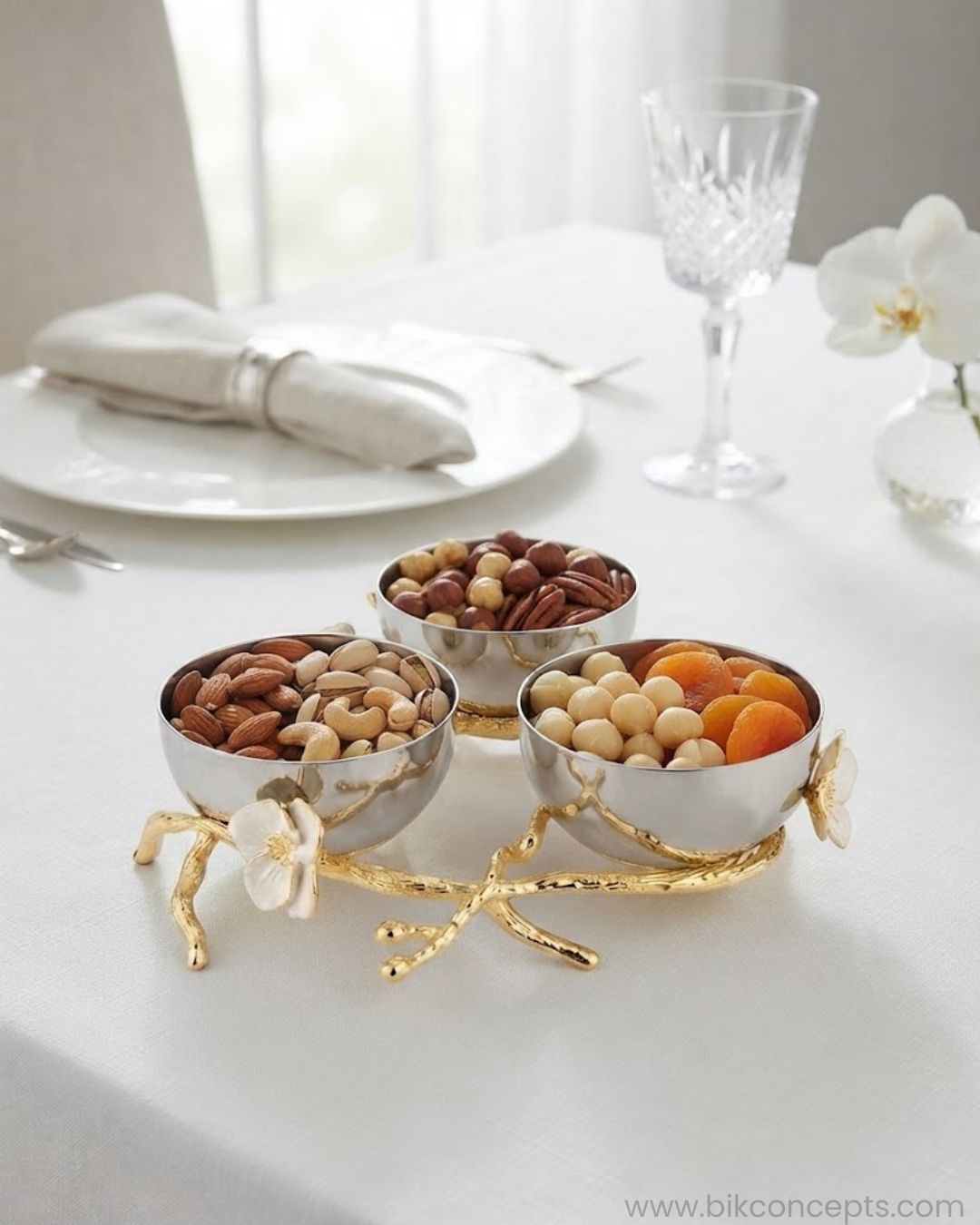 Three small metallic bowls filled with nuts and dried fruit arranged on a white tablecloth, with a gold branch-like base and white flower accents. A white plate, folded napkin, Stainless Steel Condiment Bowl Set, Luxury 3 Bowl Dip Server, Jasmine Collection Sauce Tray, High End Catering Condiment Platter USA, Silver Trio Serving Dish, Botanical Metal Snack Bowl Stand, Gold and Silver Dip Bowl Set, Designer Cocktail Garnish Tray, Luxury Wedding Tableware, Decorative 3 Tiered Condiment Dish
