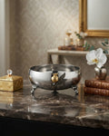 A polished silver bowl with three small feet and a gold-toned decorative accent, displayed on a dark marble surface in an elegant interior setting, 9 Inch Stainless Steel Footed Bowl, Luxury Pomegranate Centerpiece Bowl, High End Decor Bowl for Home, Botanical Metal Server for Catering, Silver Footed Serving Basin, Premium Catering Food Presentation USA, Designer Tabletop Centerpiece Vessel, Antique Gold and Silver Fruit Bowl, Luxury Wedding Tableware, Decorative Metal Bowl for Console