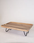 A rectangular wooden serving tray with a live-edge natural wood surface and black metal hairpin legs, Elevated Wood Serving Tray, Raised Wooden Platter with Metal Legs, Wood and Metal Serving Board, Nature Enthusiast Home Decor, Rectangular Wood Table Riser, Solid Hardwood Buffet Platter, Rustic Modern Raised Serving Tray, Black Metal Base Wood Tray, Charcuterie Platter Riser, Biophilic Kitchen Serving Accessories