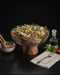 A glass pedestal salad bowl filled with a fresh mixed salad, styled on a dark tabletop with a small bowl and wooden spoon, a glass decanter, and a folded napkin with a fork nearby, Wood and Epoxy Resin Serving Bowl, Elevated Wooden Fruit Bowl, Fluted Resin Centerpiece Pedestal, Nature Enthusiast Home Accessories, Footed Wooden Snack Bowl, Resin Mosaic Decorative Bowl, Raised Timber Salad Bowl, Organic Modern Kitchen Pedestal Bowl, Artisanal Fluted Glass-Look Wood Bowl, Biophilic Home Decor Bowl