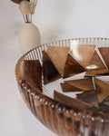 A close-up of a decorative glass bowl with triangular wooden inlays, set against a softly Wood and Epoxy Resin Serving Bowl, Elevated Wooden Fruit Bowl, Fluted Resin Centerpiece Pedestal, Nature Enthusiast Home Accessories, Footed Wooden Snack Bowl, Resin Mosaic Decorative Bowl, Raised Timber Salad Bowl, Organic Modern Kitchen Pedestal Bowl, Artisanal Fluted Glass-Look Wood Bowl, Biophilic Home Decor Bowl
