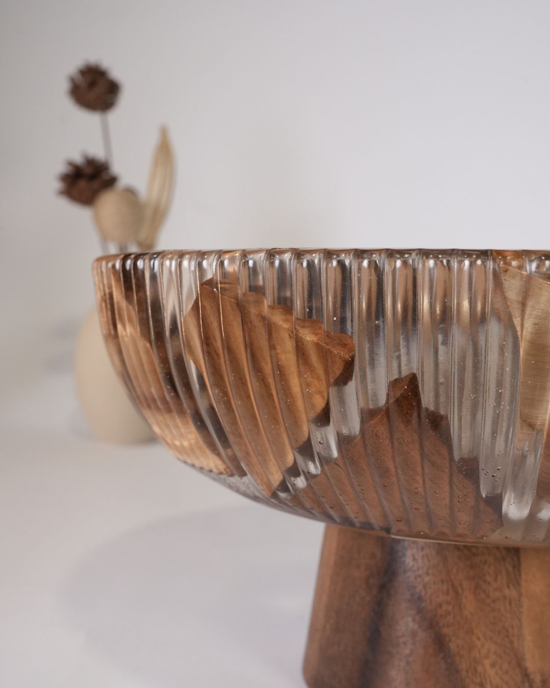 A close-up of a ribbed glass pedestal bowl with a natural wood pedestal base, set against a soft, Wood and Epoxy Resin Serving Bowl, Elevated Wooden Fruit Bowl, Fluted Resin Centerpiece Pedestal, Nature Enthusiast Home Accessories, Footed Wooden Snack Bowl, Resin Mosaic Decorative Bowl, Raised Timber Salad Bowl, Organic Modern Kitchen Pedestal Bowl, Artisanal Fluted Glass-Look Wood Bowl, Biophilic Home Decor Bowl